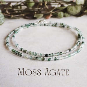 Puede incluir: Un delicado collar hecho de pequeñas cuentas redondas de ágata musgosa en tonos verdes y blancos. El collar se muestra sobre una superficie neutra con las palabras "MOSS AGATE" en una fuente decorativa.