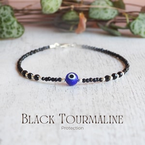 May include: A delicate bracelet featuring small, faceted black tourmaline beads interspersed with tiny silver-toned spacers. A single, round blue evil eye bead with a white and black centre is positioned at the focal point of the jewellery piece.