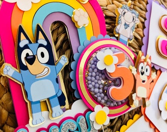 May include: A colorful cake topper featuring a rainbow, flowers, and cartoon characters. The topper includes the name "Lucy" in silver glitter letters and the number "5". The design incorporates shades of pink, blue, orange, and yellow.
