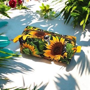 Puede incluir: Bolsa de cosméticos rectangular con un estampado vibrante de girasoles y mariposas. La bolsa tiene un tirador de cremallera amarillo y una cremallera marrón. La bolsa mide aproximadamente 20 cm de largo y 10 cm de ancho.