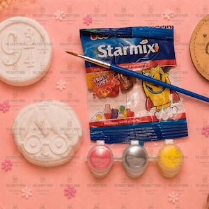 May include: A craft kit featuring two white circular plaster figures, one with the text 9 3/4 and one with a face wearing glasses. Includes a blue paintbrush, a small bag of Starmix sweets, and three small paint pots in pink, silver, and yellow.