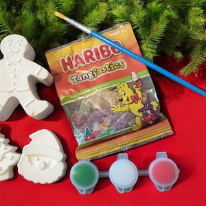 May include: A Christmas-themed craft set featuring a gingerbread man, Christmas tree, and Santa Claus figure. Includes a paintbrush, paint pots, and a bag of HARIBO Tangfastics sweets. Set on a red surface with evergreen branches.