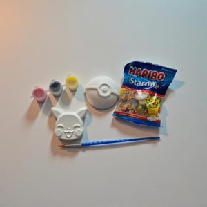 May include: A white Pikachu figure, a white Pokéball, and three small paint pots with pink, grey, and yellow paint. A blue paintbrush and a bag of Haribo Starmix sweets complete the set.