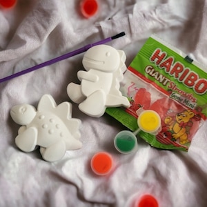 May include: Two white dinosaur figurines and a purple paintbrush are arranged on a white cloth. A small bag of Haribo Giant Straws candy and several small paint pots in yellow, green, and red are also present.