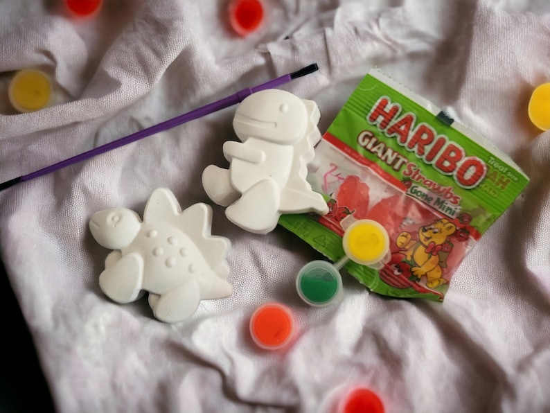 May include: Two white dinosaur figurines and a purple paintbrush are arranged on a white cloth. A small bag of Haribo Giant Straws candy and several small paint pots in yellow, green, and red are also present.