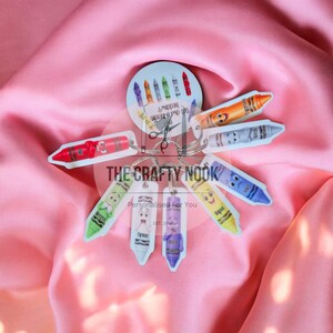 May include: A circular card featuring small crayon illustrations and the text The Crafty Nook Est. 2016, attached to a metal keyring with multiple individual crayon-shaped charms in red, orange, grey, blue, yellow, purple, white, and green colors.