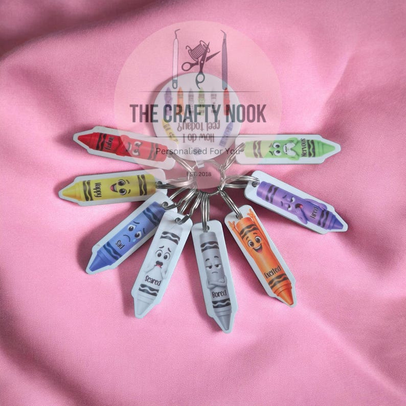 May include: A circular keychain ring holding eight individual crayon-shaped charms, each featuring a different facial expression and emotion label. The charms are red, green, yellow, purple, blue, white, grey, and orange. A central circular tag displays the text THE CRAFTY NOOK.