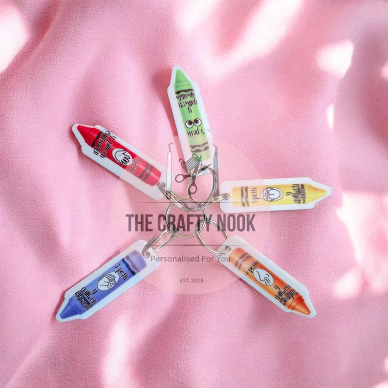 May include: A keychain featuring five crayon-shaped charms in red, green, yellow, orange, and blue. Each charm displays a unique hand sign illustration and text. The charms are attached to a central metal ring, creating a star-like arrangement.