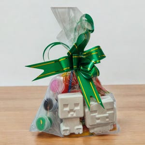 May include: A cellophane gift bag tied with a green ribbon and bow. Inside are small paint pots, sweets, and two white, blocky figures resembling characters from a popular video game.