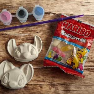 May include: A craft kit featuring two white plaster Stitch heads, three paint pots with pink, grey, and blue paint, a purple paintbrush, and a bag of Haribo Supermix sweets. The items are on a wooden surface.