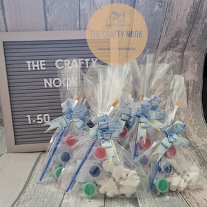 May include: Clear cellophane bags containing paint sets, each with a paintbrush, paint pots in red, blue, and green, and a white plaster figure. The bags are tied with blue and gold ribbons. The sign says "THE CRAFTY NOOK".