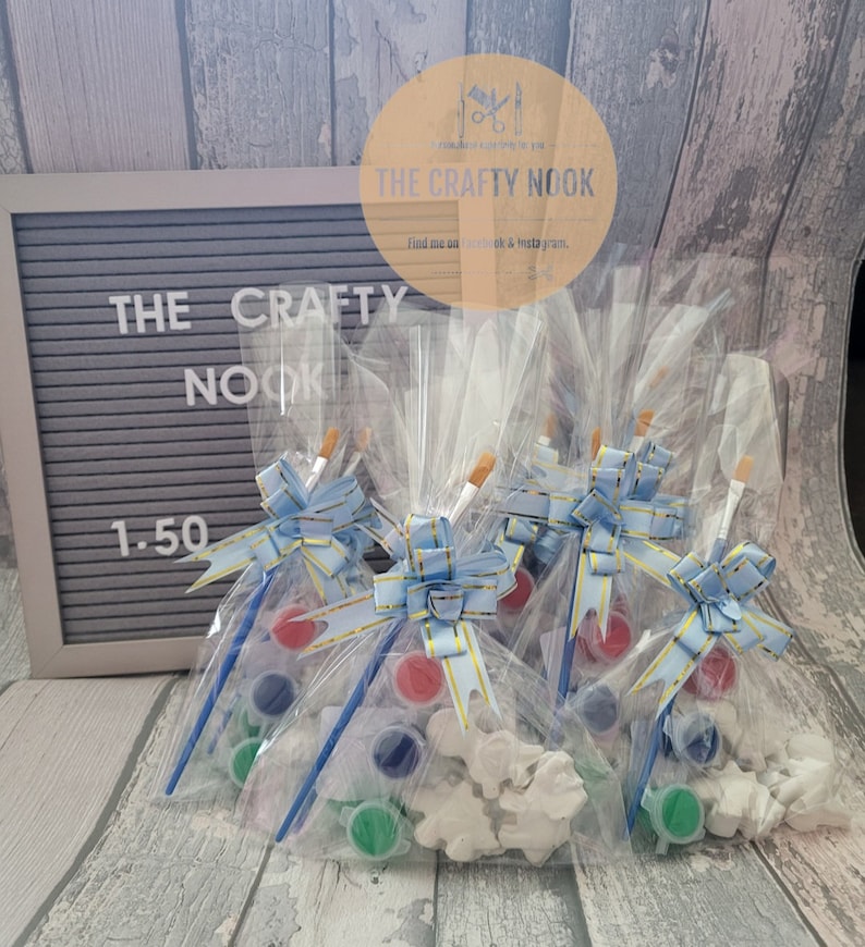 May include: Clear cellophane bags containing paint sets, each with a paintbrush, paint pots in red, blue, and green, and a white plaster figure. The bags are tied with blue and gold ribbons. The sign says "THE CRAFTY NOOK".