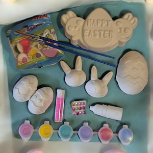 May include: An Easter craft kit with paint, brushes, and ceramic shapes. The kit includes egg and bunny shapes, paint pots, and a bag of sweets. The words "Happy Easter" are on a ceramic shape. The kit is on a blue surface.