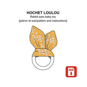 Puede incluir: Un sonajero de tela amarillo y blanco con forma de orejas de conejo con un patrón floral. El patrón incluye pequeñas flores blancas sobre un fondo amarillo. El juguete es un proyecto de bricolaje con un patrón PDF e instrucciones.