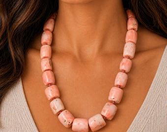 May include: A necklace featuring large, barrel-shaped beads in shades of pink and white. The beads are strung together to create a statement piece. The necklace is worn around the neck.