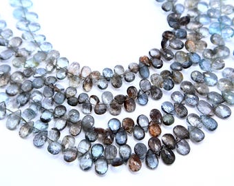 May include: Close-up of multiple strands of gemstone beads. The beads are teardrop-shaped and vary in shades of blue, gray, and brown. The beads are strung together, creating a delicate and elegant appearance. The background is white.