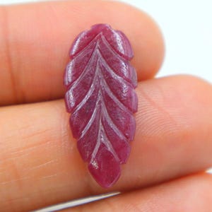 May include: A carved ruby gemstone in the shape of a leaf. The gemstone is a deep red color with detailed carved lines that mimic the veins of a leaf. The leaf shape is elongated and has a smooth, polished surface.