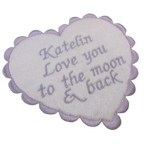May include: A white, heart-shaped plush item with a lavender scalloped edge. Lavender embroidery reads "Katelin Love you to the moon & back" across the center of the heart.