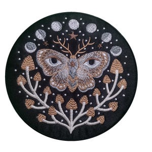 May include: Embroidered patch featuring a white moth with brown markings and large eyes, encircled by a ring of white moons and brown mushrooms on a black background.