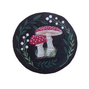 May include: A circular, black embroidered patch with a design of two red and white spotted mushrooms. The mushrooms are framed by green leaves and white berries. This patch is a decorative item.