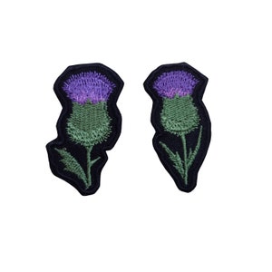 May include: Two embroidered patches featuring a thistle flower design. Each patch displays a vibrant purple bloom atop a textured green bulb and stem, all outlined with a thick black border against a solid black background.