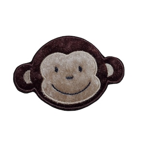May include: Embroidered patch of a smiling monkey face. The monkey's face is light tan, with dark brown eyes and a stitched mouth. The patch has a dark brown border and ear details, creating a playful design.