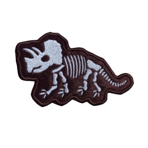 May include: A brown embroidered patch depicting a white dinosaur skeleton. The dinosaur is a triceratops with three horns and a frill.