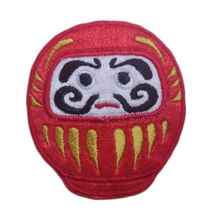 May include: Embroidered red Daruma doll patch with a white face, black facial features, and gold accents. The Daruma doll is a traditional Japanese good luck charm, often used for goal setting and motivation.