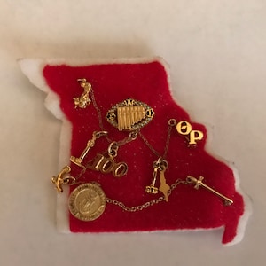 May include: A red felt Missouri state outline with white trim, adorned with gold-colored charms and chains. Charms include a musical instrument, anchors, and the number "100".