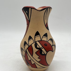 以下が含まれることがあります： フレアリムの小さなタン色のセラミックピッチャー。ピッチャーは、鳥のようなモチーフを含む、赤、黒、白の幾何学模様の様式化されたデザインで飾られています。リムは赤く塗られています。
