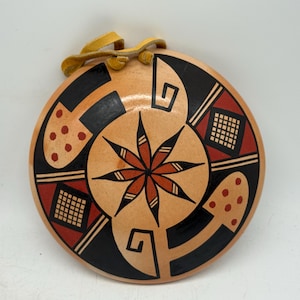 May include: Round, tan-colored decorative plate with a central starburst design in red and black. The plate features geometric patterns and a leather strap for hanging. The design includes black shapes, red accents, and small red dots.