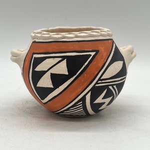 Puede incluir: Una pequeña olla de cerámica pintada a mano con un diseño geométrico en negro, blanco y naranja. La olla tiene una forma redondeada y dos pequeñas asas.