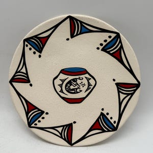 Könnte beinhalten: Runder, cremefarbener Keramikteller mit einem zentralen Design und einem sich wiederholenden geometrischen Muster. In der Mitte befindet sich ein Topf mit einer Figur. Das geometrische Muster ist schwarz mit roten und blauen Akzenten.