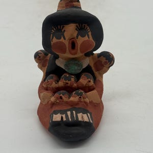 May include: A hand-painted ceramic figurine with a stylized face, large eyes, and an open mouth. The figure wears a black hat and a turquoise necklace. The base is reddish-brown, and the design is whimsical. The figurine is likely a decorative item.