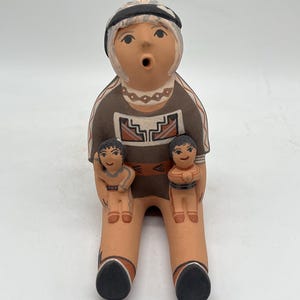 May include: A handcrafted ceramic figurine depicting a seated figure with two smaller figures. The main figure wears a brown top with a geometric design and a black and white head covering. The smaller figures are held on the main figure's legs.