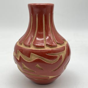 May include: A handcrafted red ceramic vase with a rounded body and a narrow neck. The vase features carved geometric patterns in a lighter tan color, creating a contrasting design. The vase is approximately 6 inches tall, perfect for home decor.