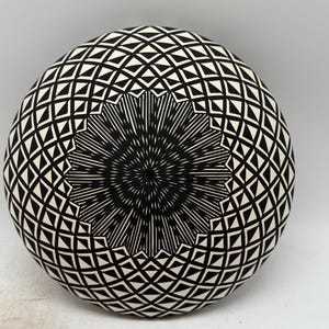 May include: A round, black and white ceramic bowl with a geometric pattern. The design features a central starburst motif surrounded by diamond and triangle shapes. The bowl's surface is smooth and the overall aesthetic is modern.