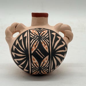 Puede incluir: Un pequeño jarrón de cerámica pintado a mano con una base de color beige claro y un borde rojo oscuro. El jarrón presenta dos asas de bucle y un diseño central de patrones geométricos negros y naranjas.