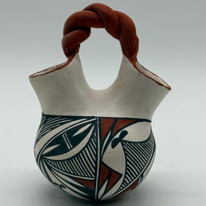 May include: A ceramic vessel with two spouts and a central twisted handle in a terracotta colour. The body is white with black and terracotta geometric patterns, featuring fine line work and angular shapes across the rounded base of the object.