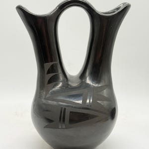 May include: A black pottery wedding vase with a distinctive handle and flared openings. The vase features etched geometric designs, including triangles and lines, adding a decorative element to the traditional form. The vase is a handcrafted piece.