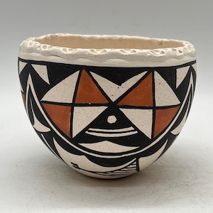 Puede incluir: Un pequeño cuenco de cerámica blanca con un diseño geométrico en negro y naranja. El cuenco tiene un borde festoneado y está decorado con un patrón repetitivo de triángulos y diamantes.