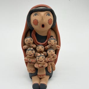 Puede incluir: Una figura de cuentacuentos de terracota que presenta una figura central grande con cabello negro y mejillas rojas, rodeada por cinco figuras más pequeñas. Las figuras están pintadas con detalles en negro y rojo, y la figura central lleva un chal rojo.