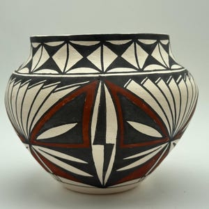 Może przedstawiać: Zaokrąglony ceramiczny garnek o kremowej podstawie z ręcznie malowanymi geometrycznymi wzorami w kolorze czarnym i terakotowej czerwieni. Projekt zawiera pas trójkątów wokół krawędzi oraz duże, symetryczne motywy w kształcie wachlarzy.