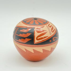 Könnte beinhalten: Ein rundes, handbemaltes Keramikgefäß mit beigefarbener Basis und komplexen geometrischen Mustern in Schwarz und Terrakotta-Rot. Die Oberseite zeigt ein kreisförmiges Blumenmotiv, während die Seiten mit treppenartigen und geschwungenen Formen verziert sind.