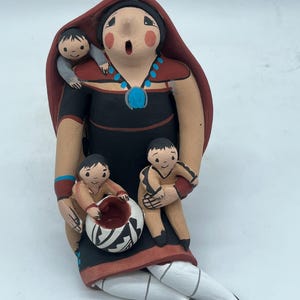 Puede incluir: Una figurita de arcilla pintada a mano de una mujer con tres niños. La mujer lleva una chal roja y un vestido negro con joyas turquesa. Está sosteniendo una pequeña olla de arcilla en su regazo. Los niños están sentados en su regazo y hombros.