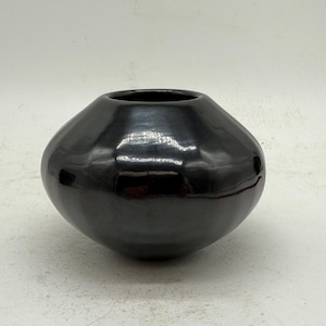 May include: A black, rounded ceramic vase with a wide opening. The vase has a glossy finish and a smooth, curved shape. The dark color and simple design make it a versatile decorative piece for any home.