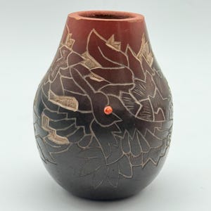 May include: A rounded vase with a tapered neck, featuring a dark brown to reddish-brown gradient finish. The surface is decorated with carved floral patterns and a single small, bright orange circular accent embedded on the front side.