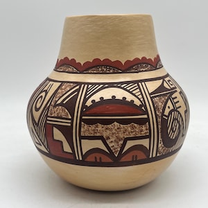 May include: A beige ceramic vase with a rounded body and a narrower neck. The vase is adorned with geometric patterns in brown, red, and cream colors. The design includes lines, shapes, and a scalloped border near the top. A decorative piece.