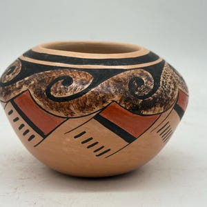 May include: A hand-painted, round ceramic pot with a wide opening. The pot features a tan base with black, brown, and red geometric designs. The upper portion has black bands and swirling patterns. The pot is likely used for decorative purposes.