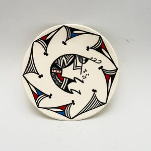 May include: A round ceramic plate with an off-white base color featuring a central black, red, and blue geometric design. The pattern consists of a spiraling, abstract motif surrounded by a series of repeating triangular shapes along the edge.
